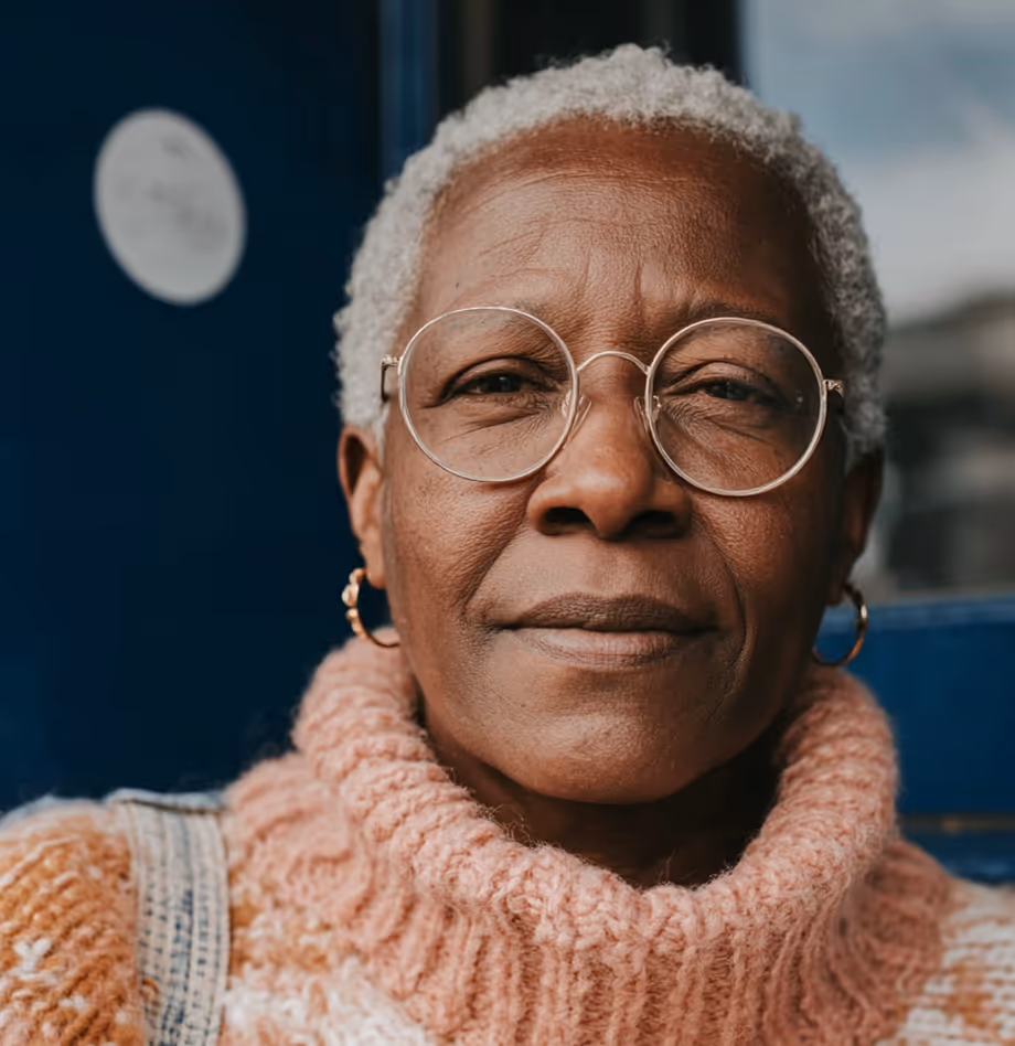 Portrait d'une femme âgée portant des lunettes rondes et un pull à col roulé rose tricoté.