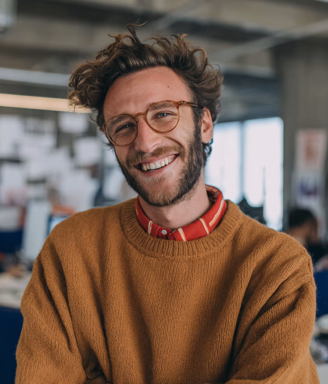 Jeune homme souriant avec lunettes, pull marron et chemise rouge à rayures en bureau moderne.