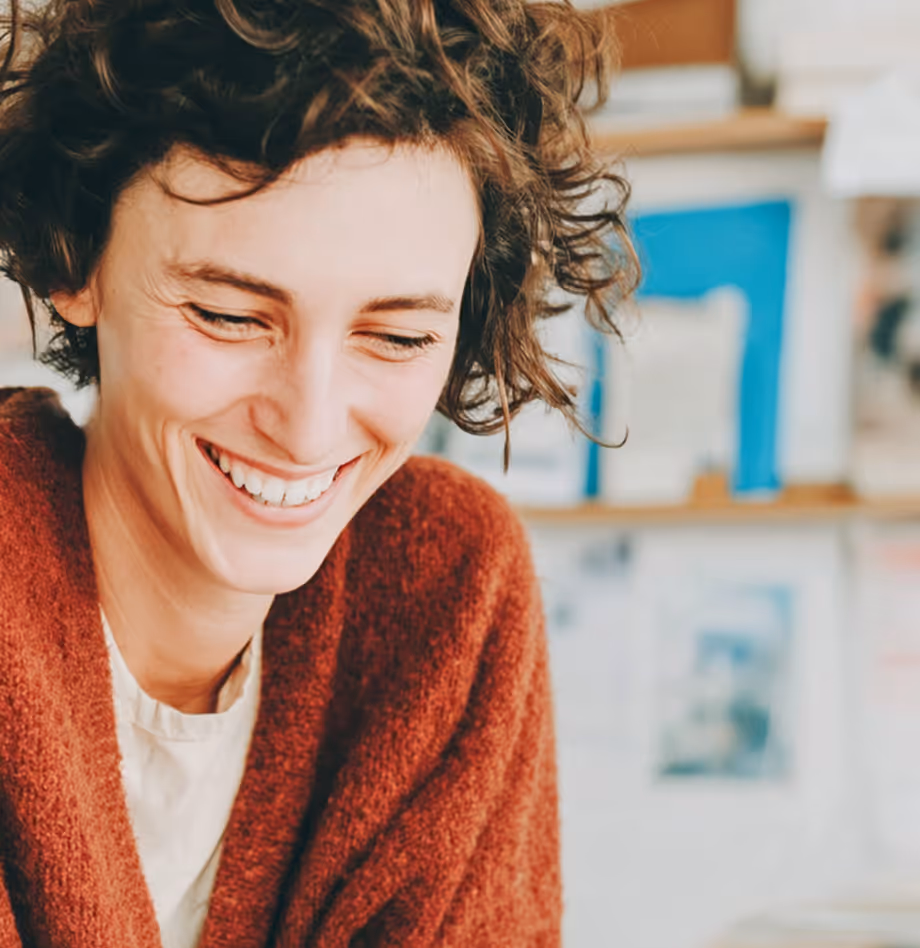 Femme souriante aux cheveux bouclés portant un pull rouge brique dans un intérieur flou.
