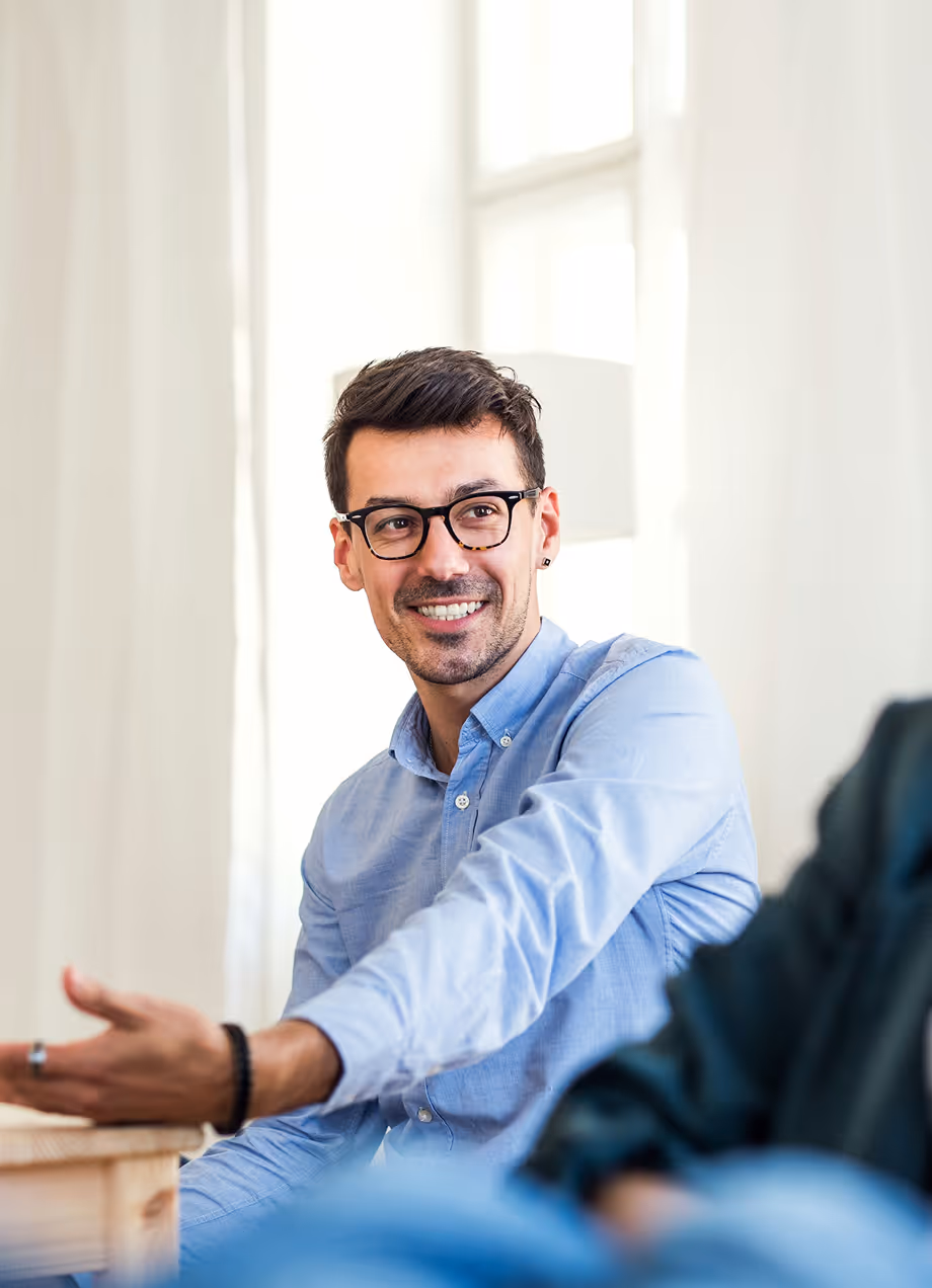 Un homme souriant portant une chemise bleue et des lunettes, gesticulant pendant une conversation.