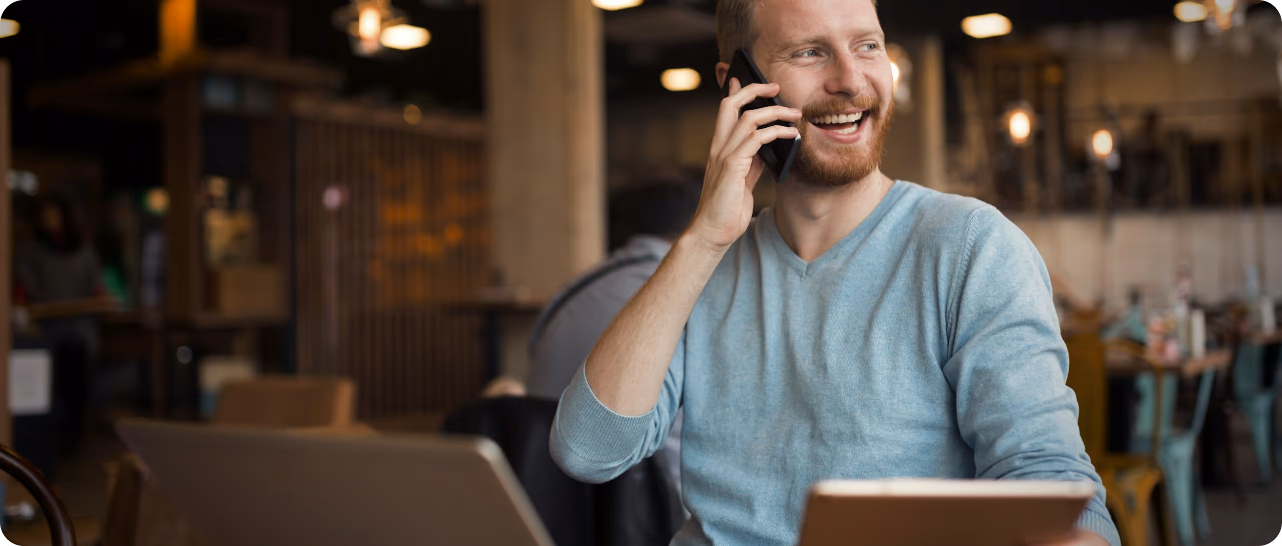 Homme souriant parlant au téléphone en travaillant avec un ordinateur portable dans un café.