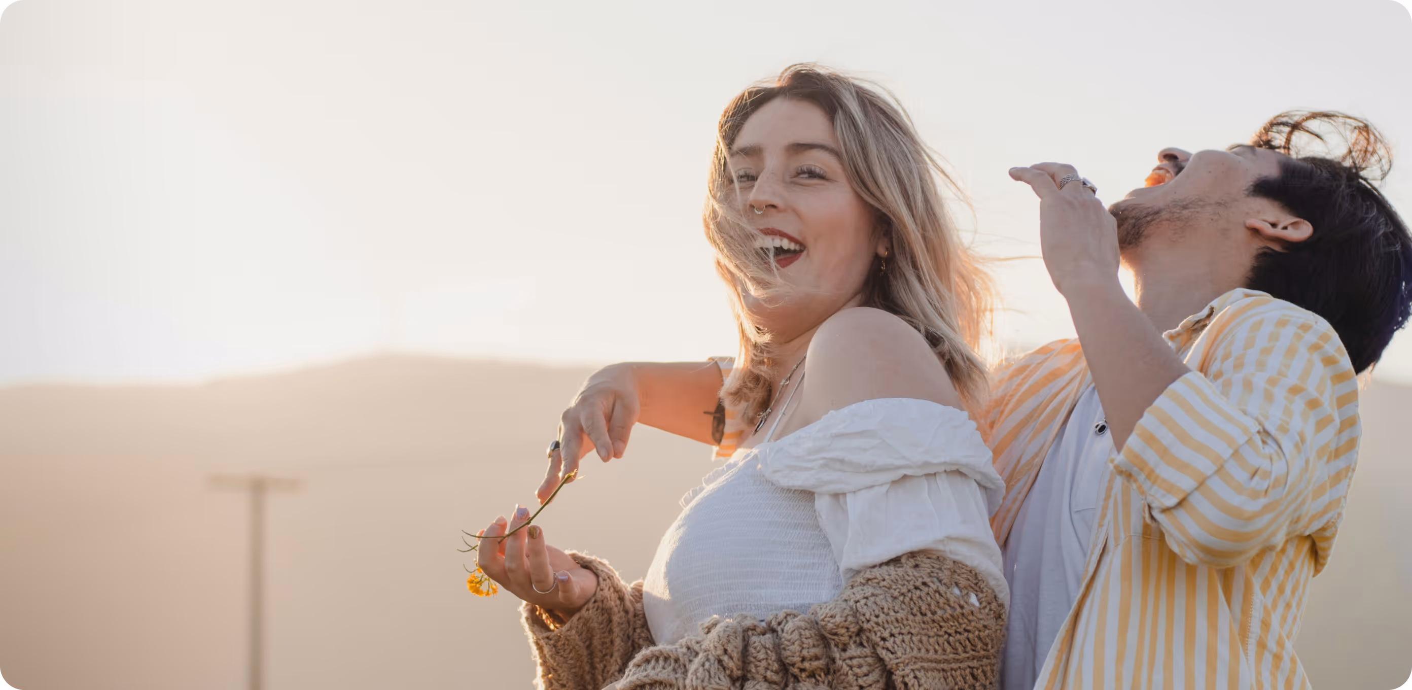 Un couple rit joyeusement dehors au coucher du soleil, une femme tient une petite fleur jaune.