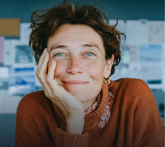 Femme souriante aux yeux bleus posant sa tête sur sa main dans un pull marron.