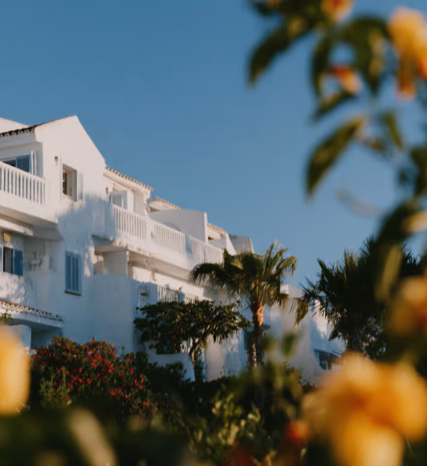 Bâtiment blanc avec balcons, plantes vertes, arbres et ciel bleu clair en arrière-plan.