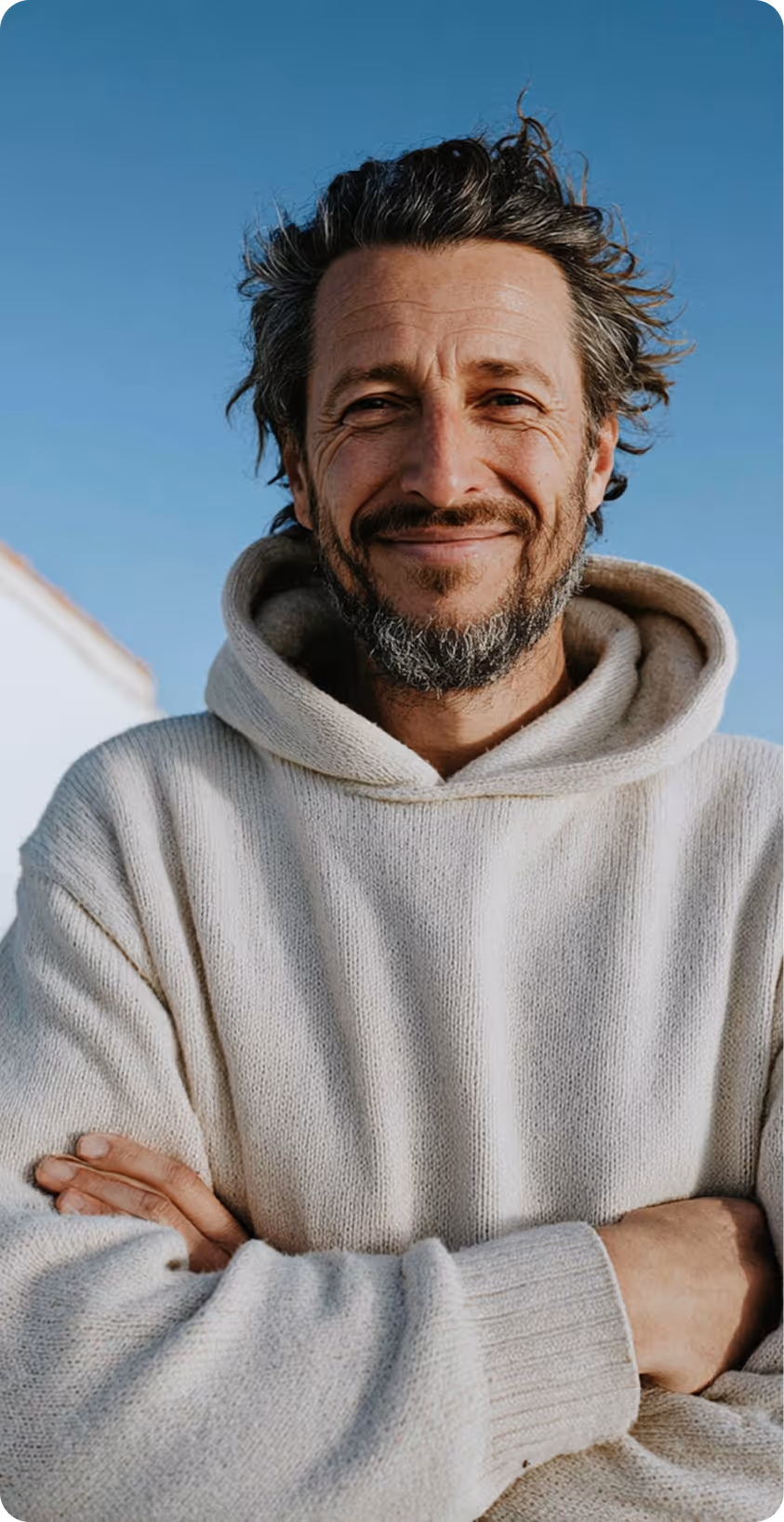 Homme souriant avec une barbe grise, bras croisés, en extérieur.