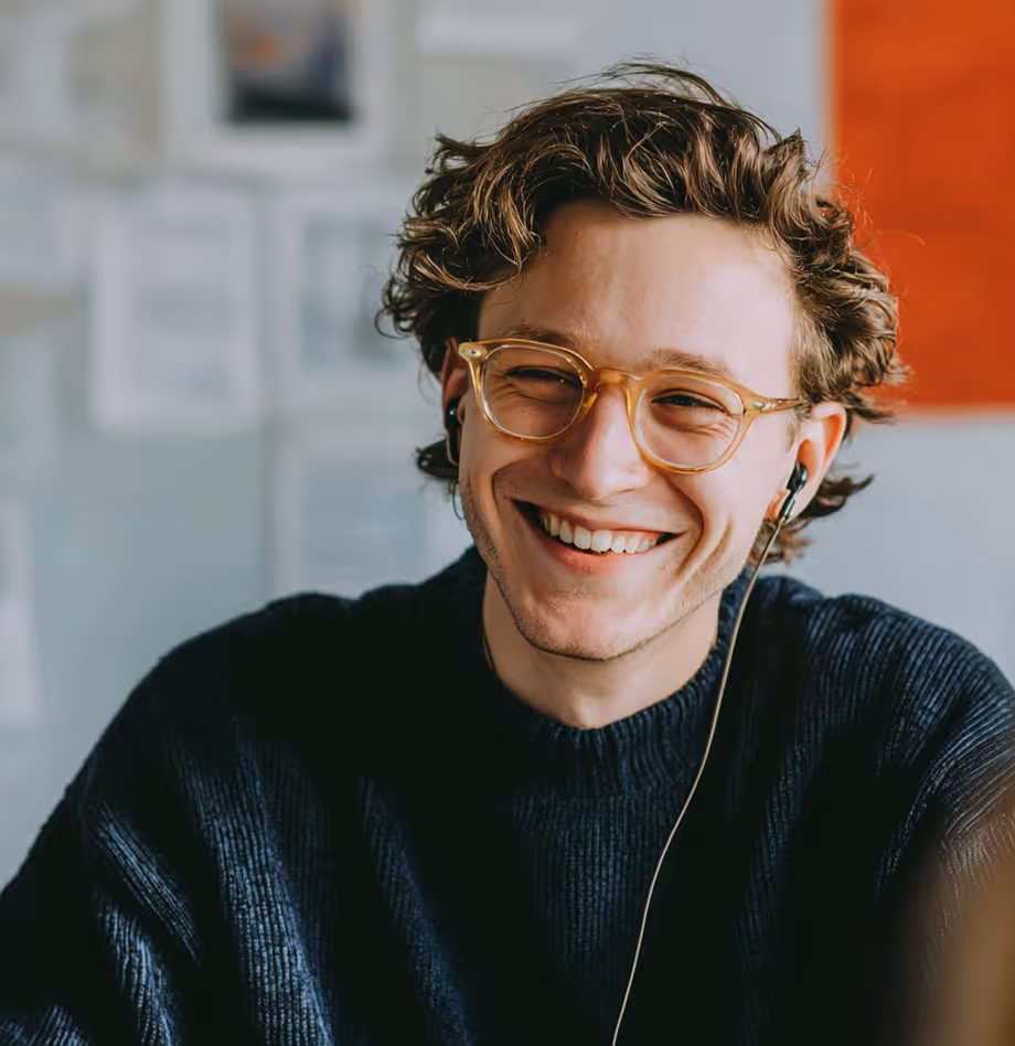 Jeune homme souriant avec des lunettes transparentes et des écouteurs, portant un pull noir, assis devant un mur avec des affiches floues.
