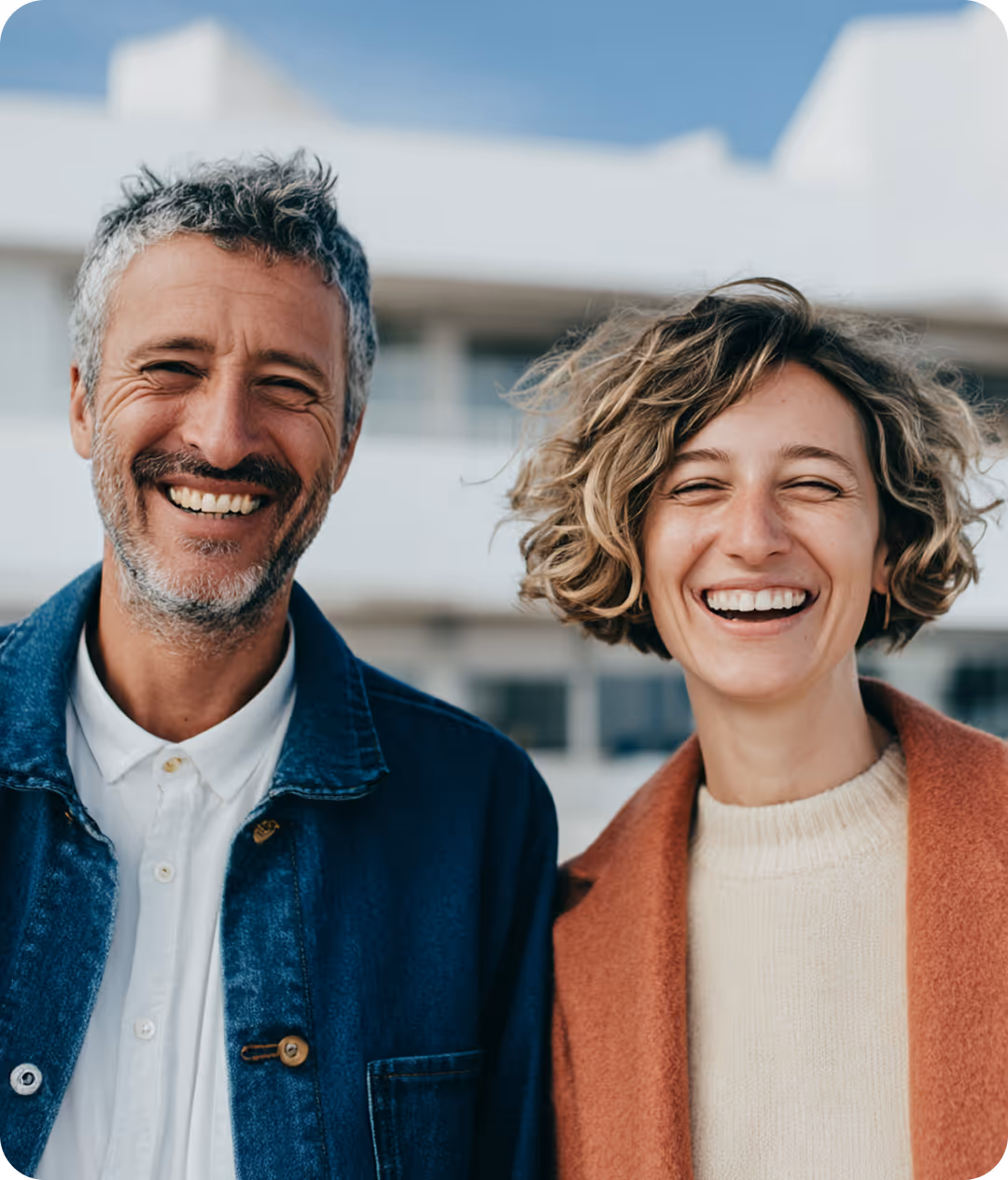 Portrait d'un homme et d'une femme souriants côte à côte, l'homme porte une veste en jean bleue et une chemise blanche, la femme porte un pull beige et un manteau marron clair, en arrière-plan un bâtiment flou sous un ciel bleu.