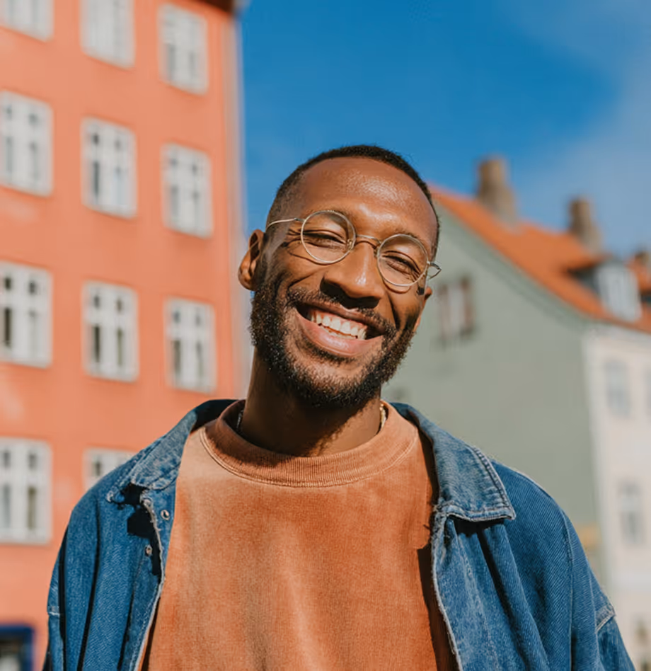 Un homme souriant portant des lunettes rondes, un sweat-shirt marron clair et une veste en jean, devant des bâtiments résidentiels colorés sous un ciel bleu.