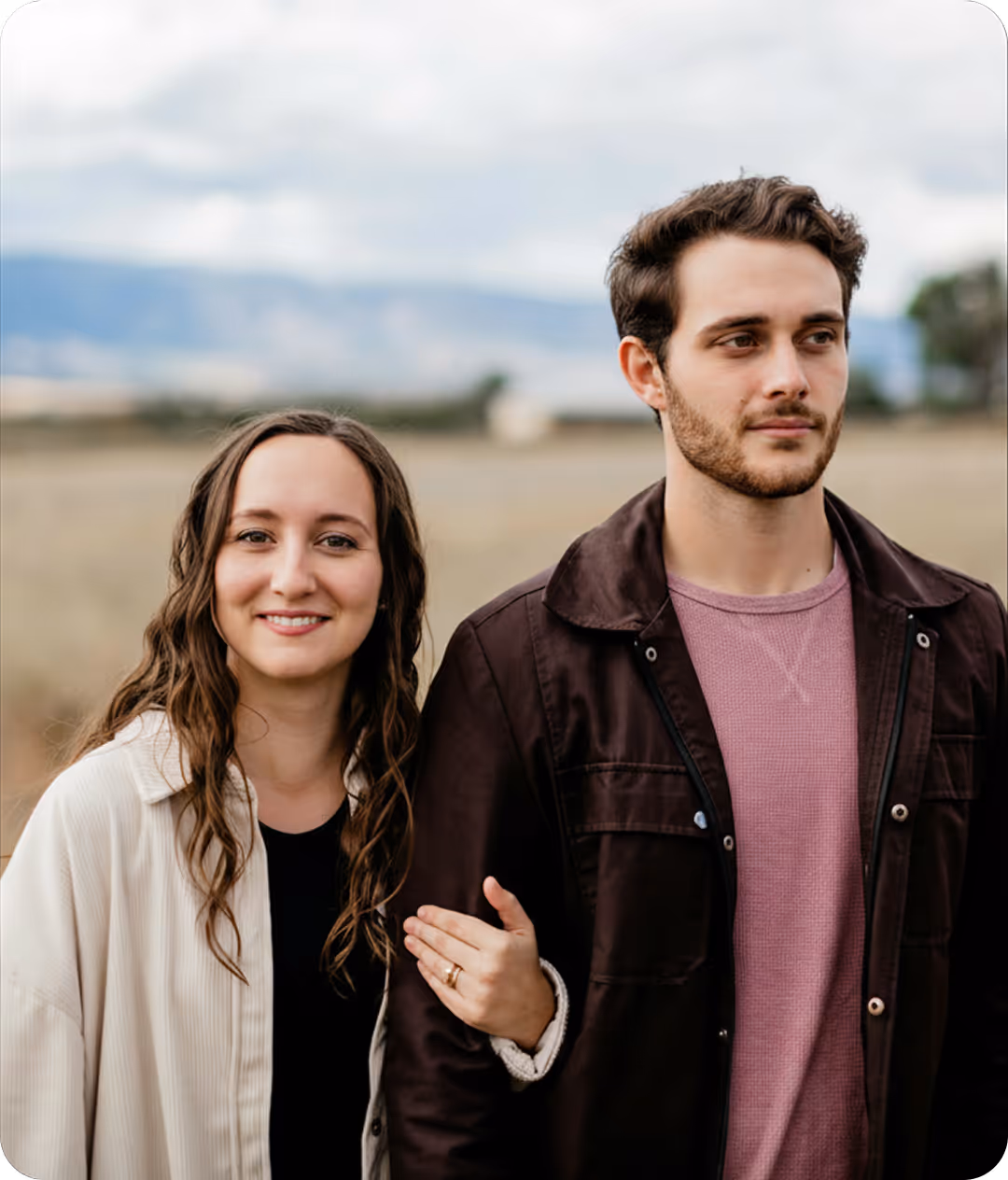 Un couple debout dans un champ, la femme souriant et tenant le bras de l'homme qui regarde au loin.