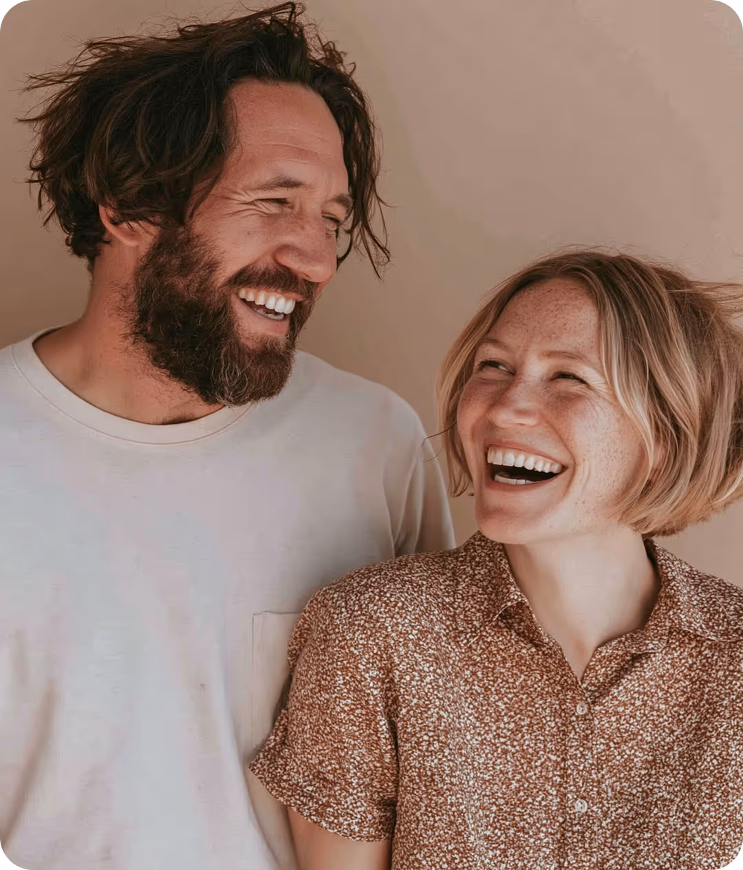 Un homme avec une barbe et des cheveux bouclés regarde et rit avec une femme aux cheveux blonds courts qui porte une chemise à motifs, tous deux souriants et face à face.