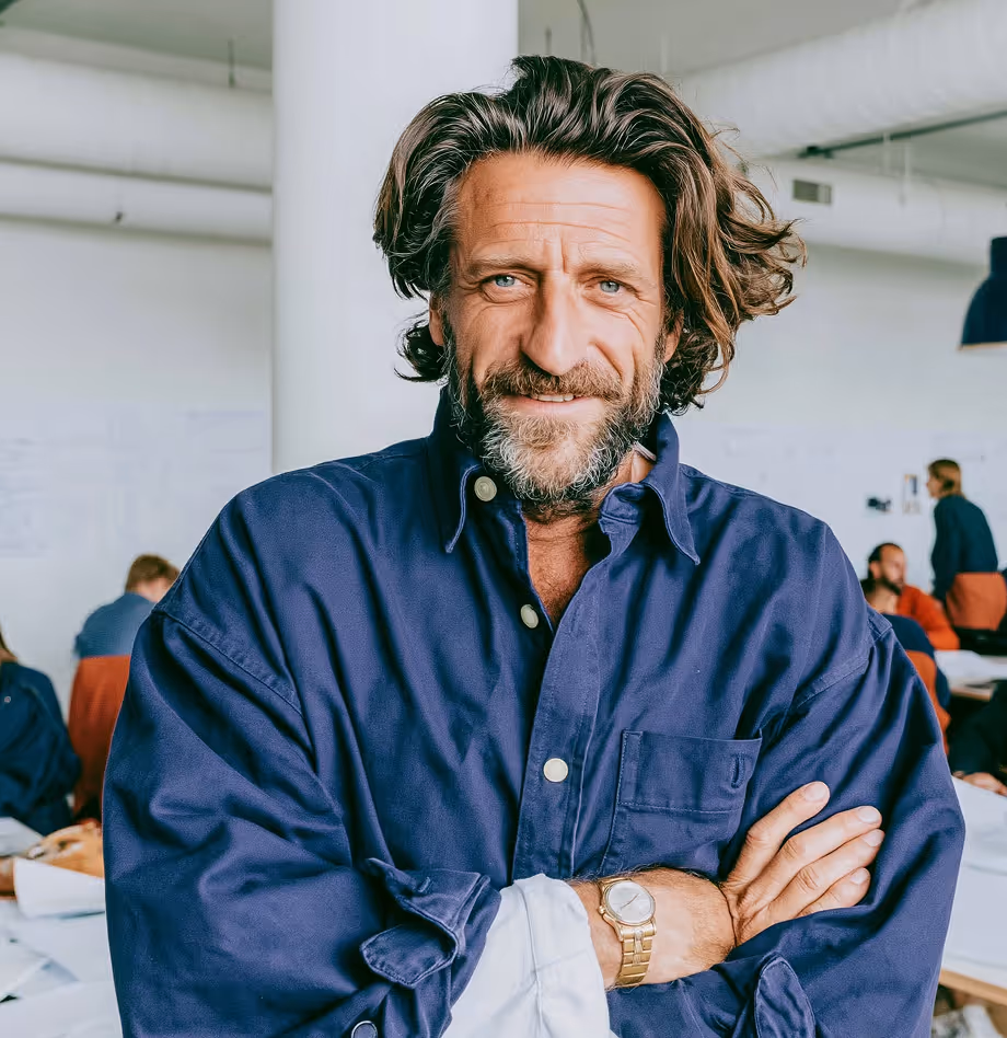 Homme souriant aux cheveux longs et barbe, portant une chemise bleue et une montre bracelet dorée, bras croisés, debout dans un bureau avec plusieurs personnes en arrière-plan.