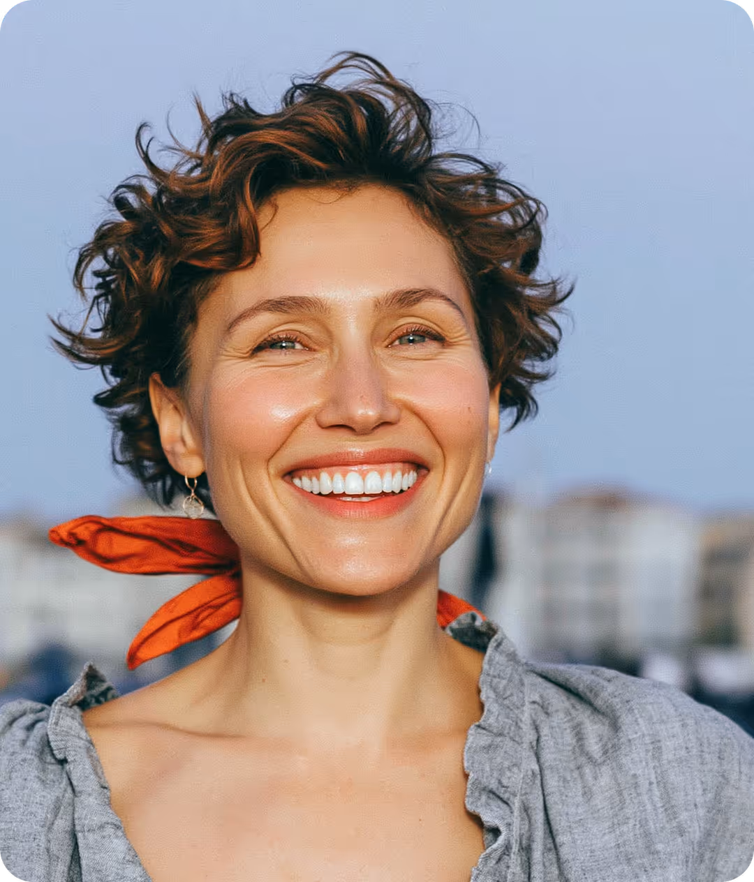 Femme souriante avec cheveux bouclés courts, portant un foulard rouge autour du cou et un haut gris, fond urbain flou.