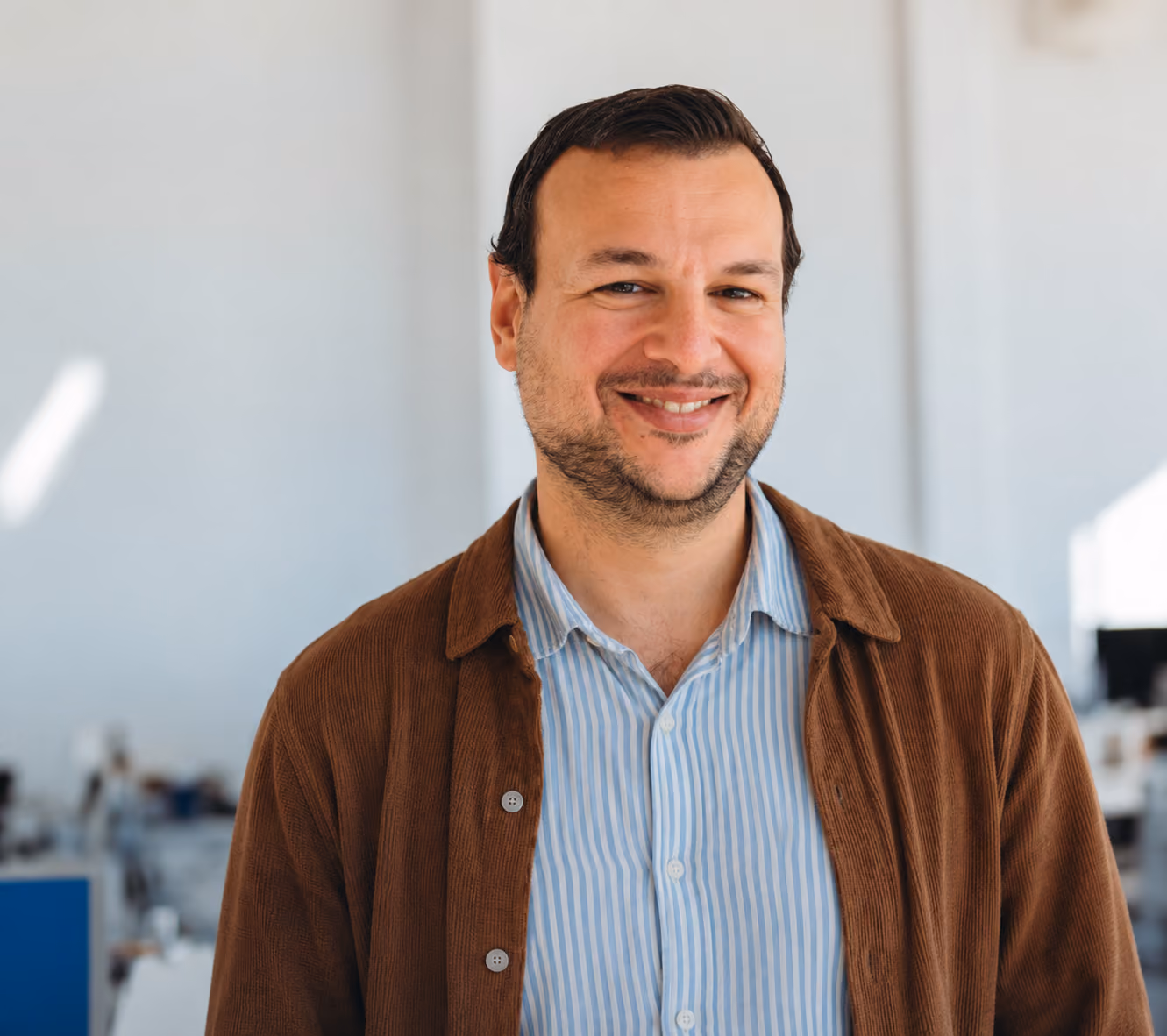 Homme souriant portant une chemise rayée bleue claire sous une veste marron, avec un arrière-plan de bureau flou.