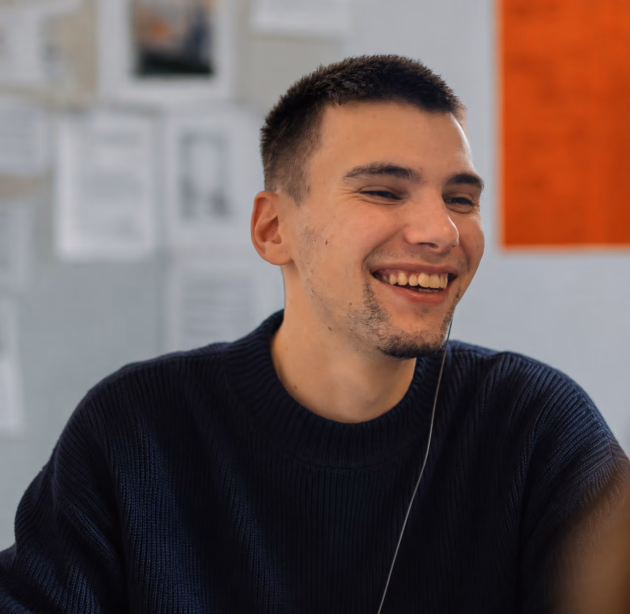 Jeune homme souriant avec un pull noir, regardant légèrement sur le côté dans un environnement intérieur flou.