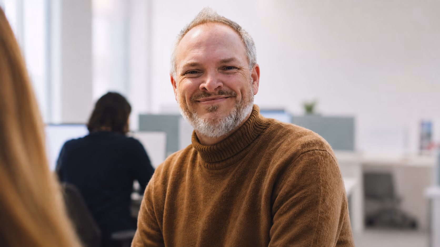 Homme souriant avec un pull à col roulé marron clair dans un bureau lumineux avec collègues en arrière-plan.