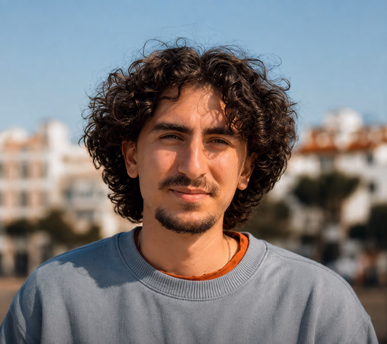 Portrait d'un homme jeune aux cheveux bouclés noirs et barbe fine, portant un pull gris clair avec un t-shirt orange en dessous, devant un fond flou de bâtiments et ciel bleu.