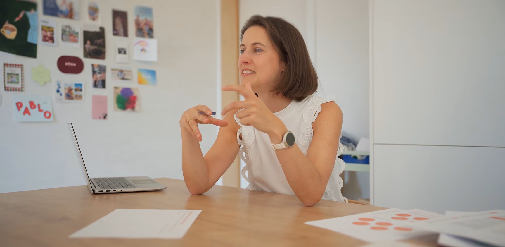 Vrouw in een witte mouwloze top die aan tafel zit en praat, met een open laptop en papieren verspreid op de tafel, en een muur met foto's en tekeningen op de achtergrond.