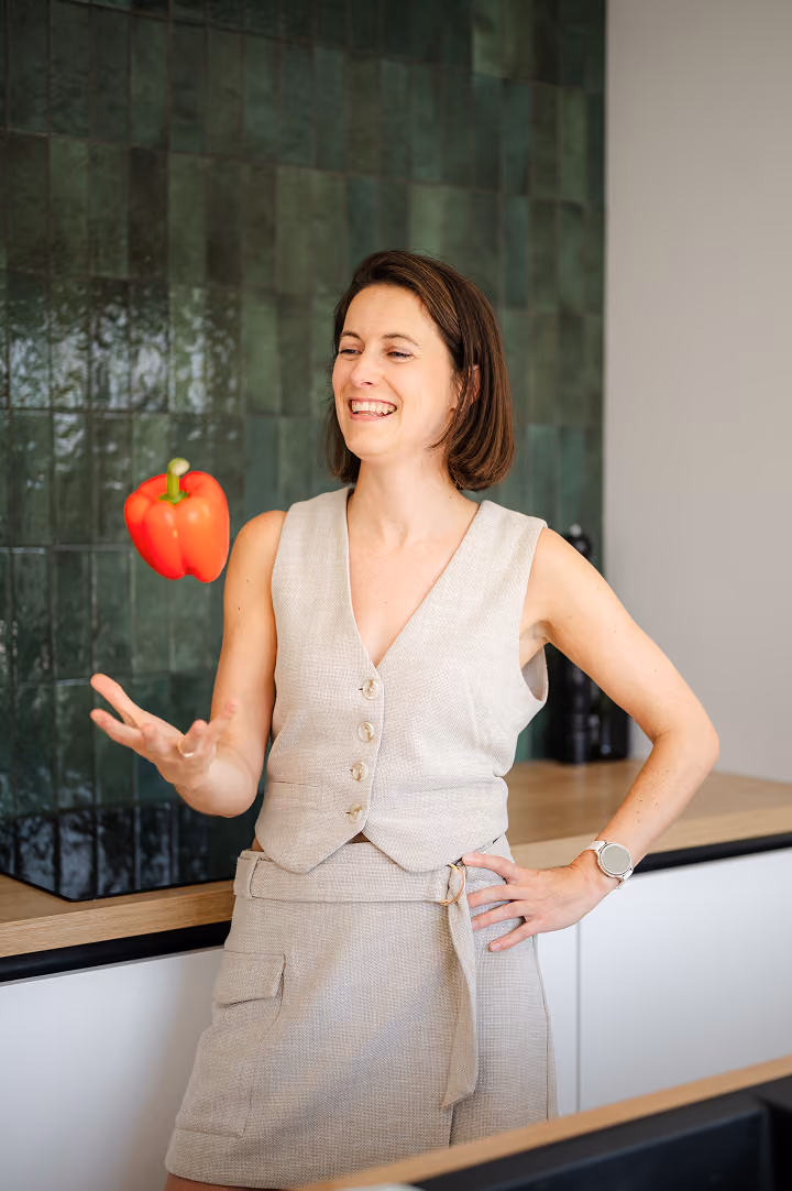 Een vrouw lacht terwijl ze een rode paprika omhoog gooit in een moderne keuken met groene tegels aan de muur.