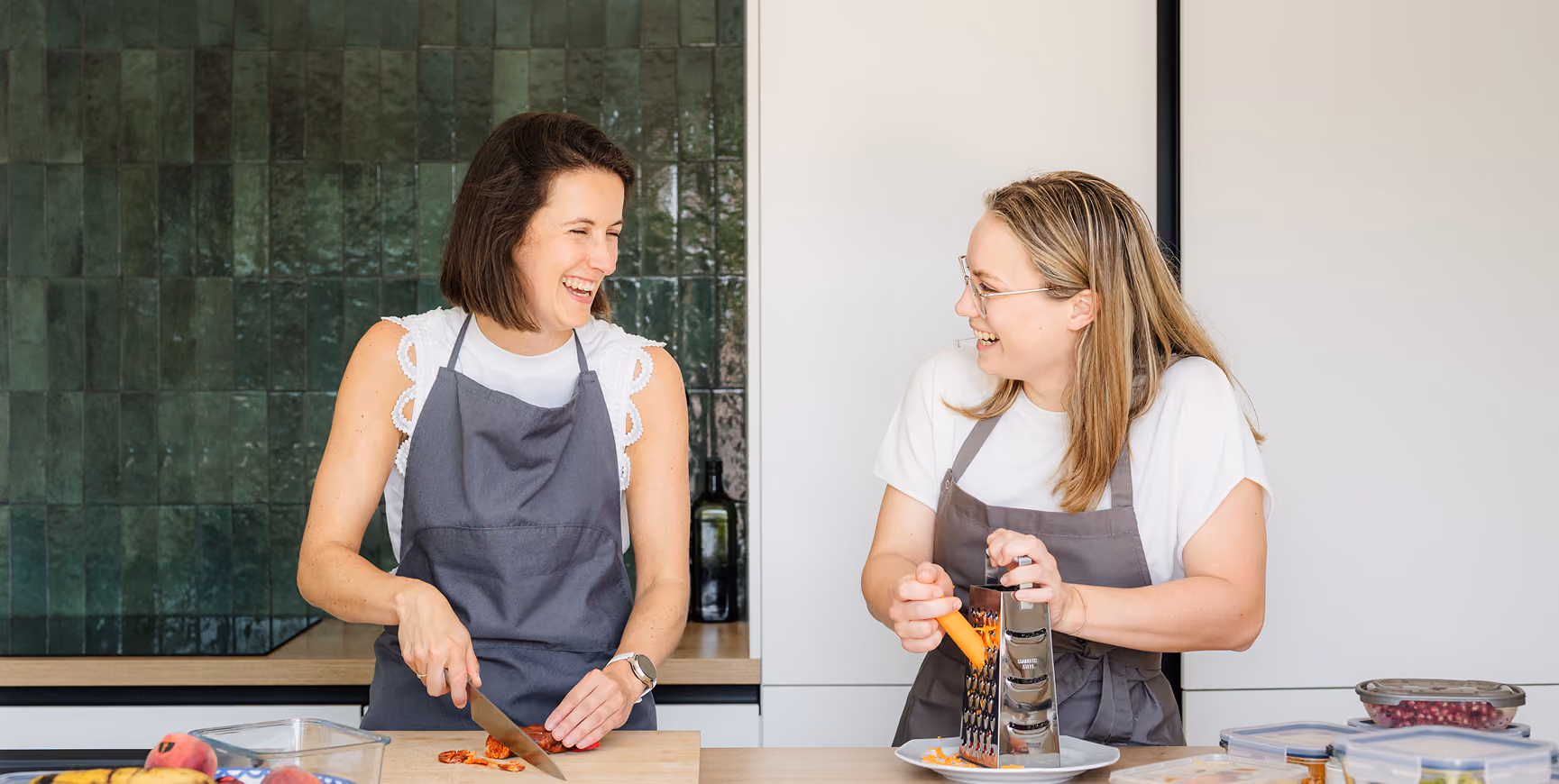 Twee vrouwen in een moderne keuken lachen en bereiden samen voedsel; de linker vrouw snijdt groenten op een snijplank en de rechter vrouw raspt wortel met een rasp boven een bord.