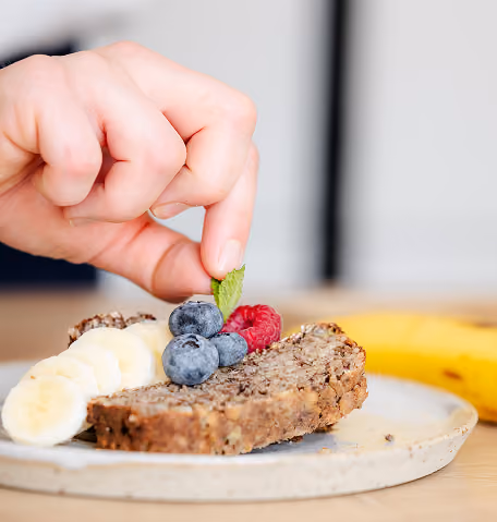 Hand die een muntblad op een plak zelfgemaakt bananenbrood legt, naast plakjes banaan en bessen op een bord.