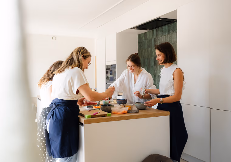 Vier vrouwen staan rond een keuken-eiland en bereiden samen een maaltijd met verschillende ingrediënten in kommen en containers.