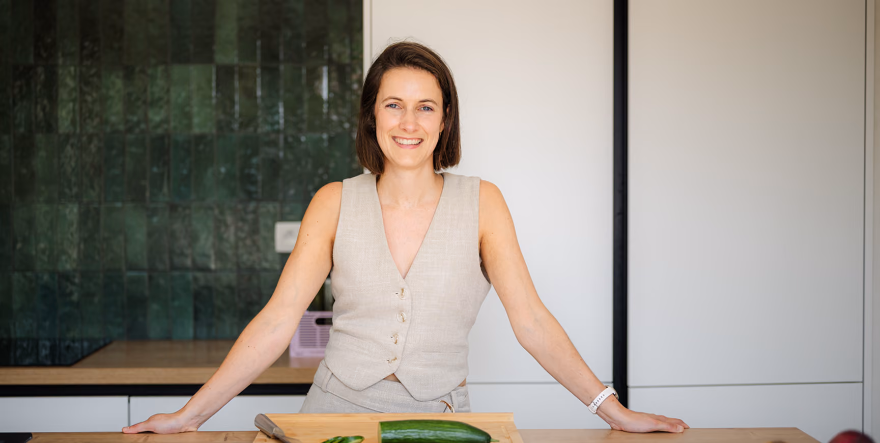 Vrouw met kort bruin haar en een lichte mouwloze blouse die lacht terwijl ze achter een keukenblad staat met een komkommer en een mes voor zich.