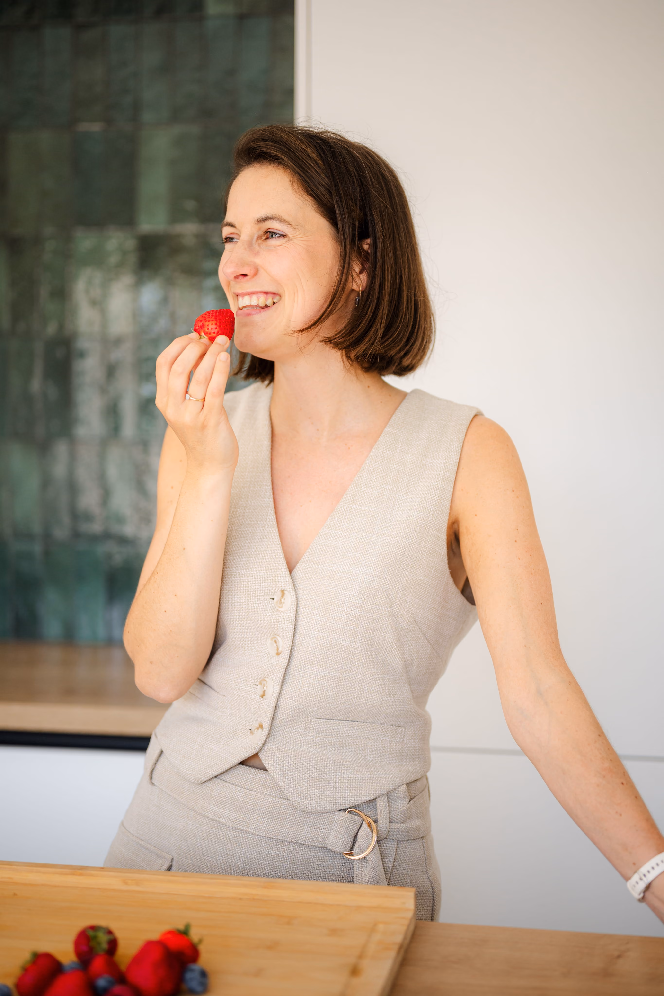Vrouw met kort bruin haar en een lichtgrijze mouwloze outfit staat in een keuken en eet een aardbei met een glimlach.
