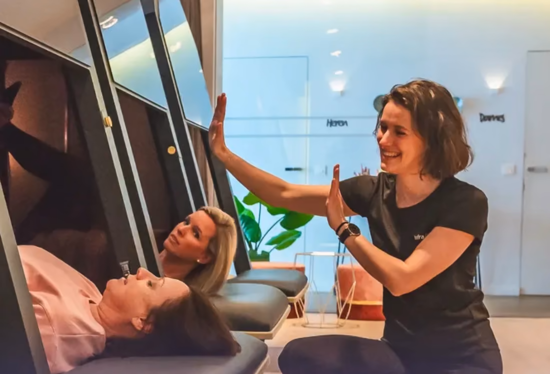 Een vrouw in zwarte kleding lacht terwijl ze haar hand omhoog houdt tegenover twee vrouwen die in een warmtecabine liggen met hun hoofd omhoog gericht