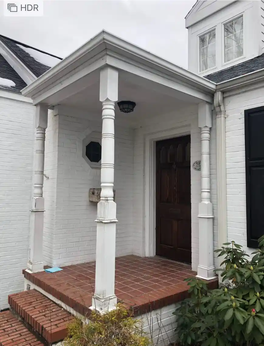 Covered porch with white columns, brick steps, a dark wooden door, and plants nearby.