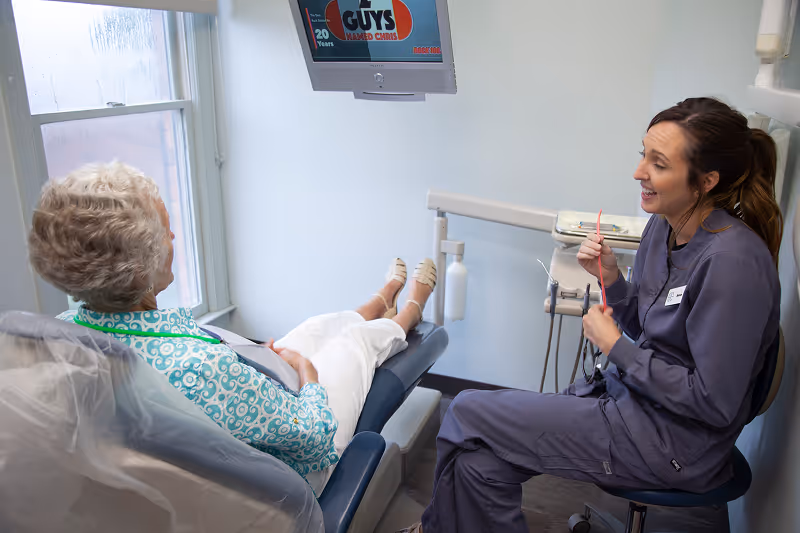 Dental hygienist talks and smiles with elderly female patient reclining in dental chair.