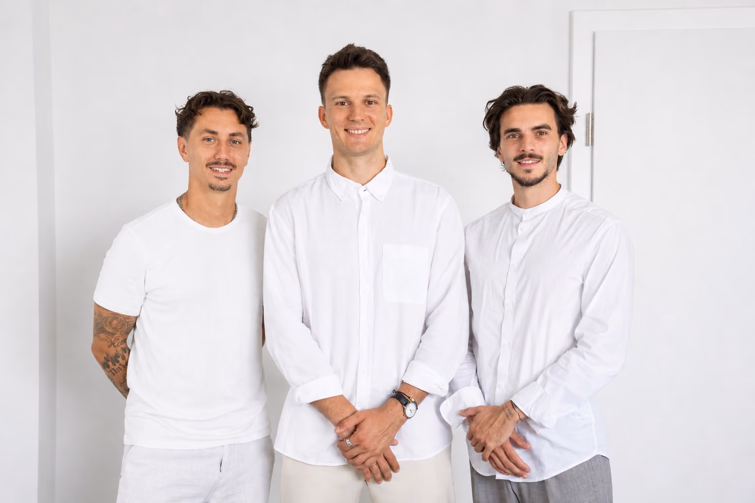 Drei junge Männer stehen vor weißer Wand, tragen weiße Oberteile und lächeln in die Kamera.