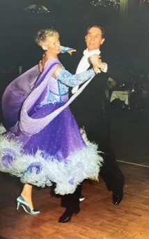 Ein Paar tanzt elegant, die Frau trägt ein lilafarbenes Ballkleid mit Federn am Saum.