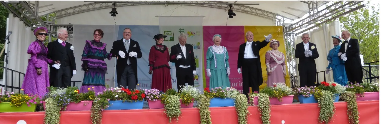 Zwölf Personen in historischer Kleidung stehen auf einer Bühne mit Blumenarrangements davor.