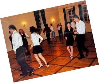 Pärchen tanzen in einem Saal mit Holzfußboden und gelber Wandbeleuchtung.