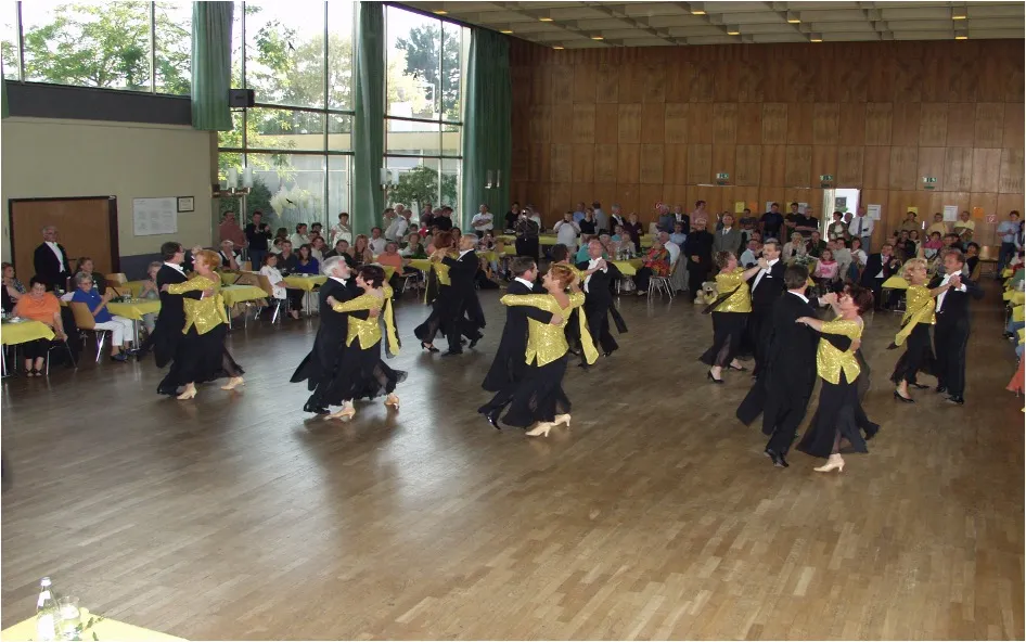Mehrere Paare tanzen in einem großen Raum vor Publikum, Frauen tragen goldene Oberteile, Männer schwarze Anzüge.