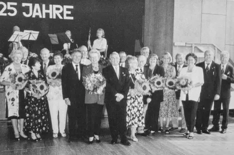 Schwarz-weiß-Foto von einer Gruppe älterer Menschen mit Sonnenblumen vor einer Bühne mit Schrift '25 JAHRE'.