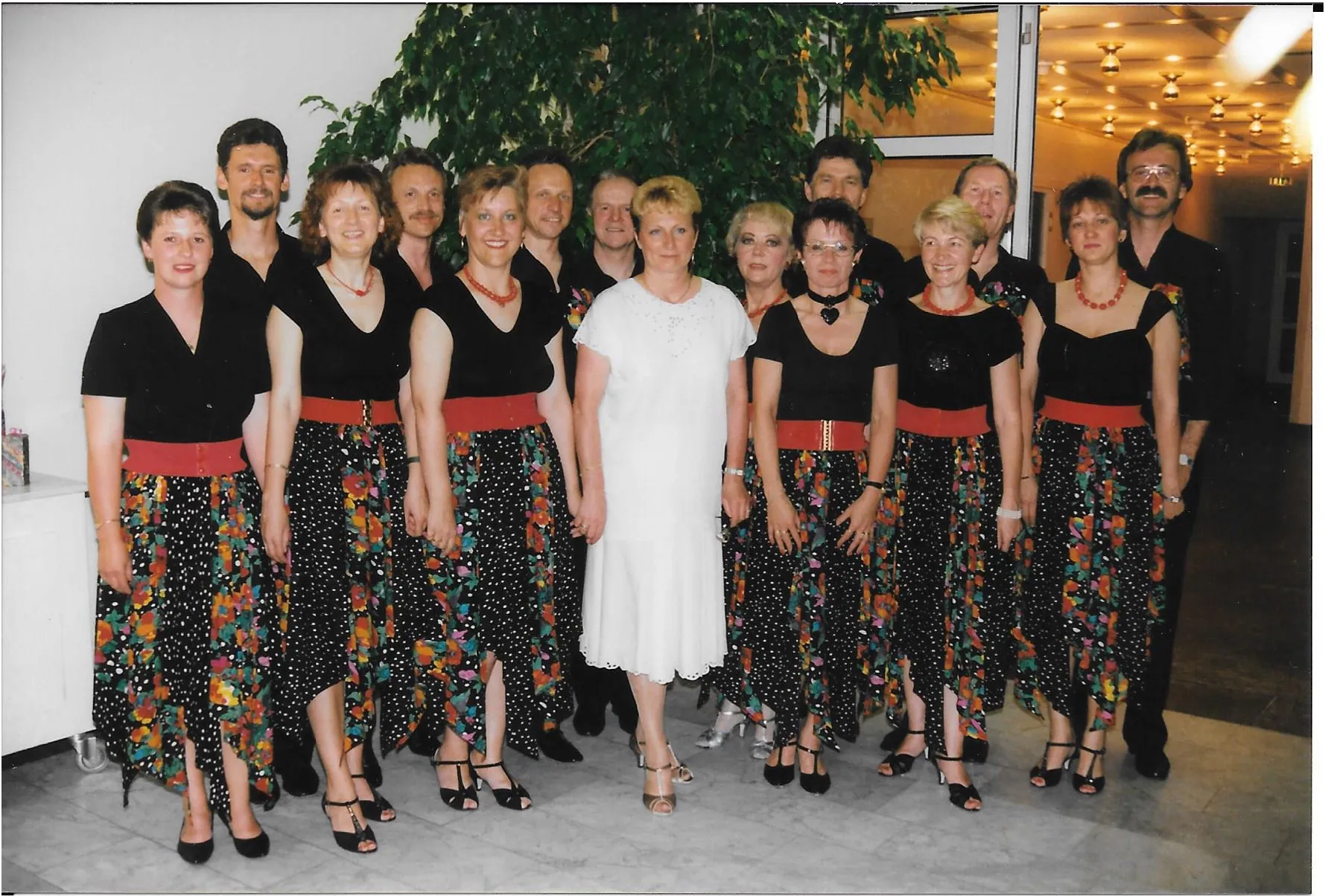 Gruppe von Erwachsenen in bunten Röcken mit roten Gürteln und einem weißen Kleid, stehend im Innenraum.