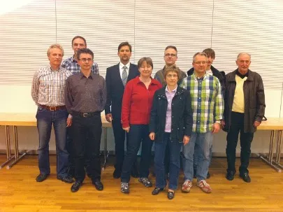 Neun Personen in legerer Kleidung stehen in zwei Reihen in einem Raum mit Holzboden.