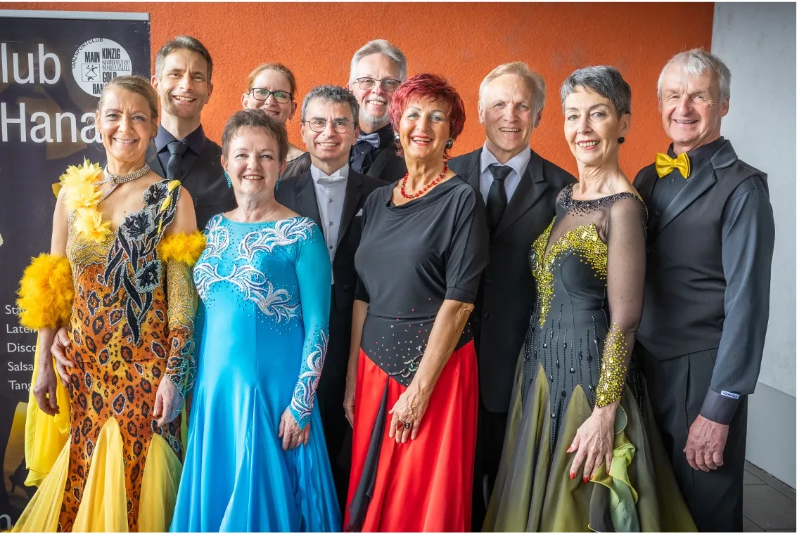 Zehn Personen lächeln, tragen elegante Tanzkleidung, vor einer orangen Wand.
