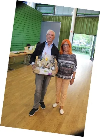 Älteres Paar steht in einem Raum, der Mann hält einen Geschenkkorb mit gedeckten Produkten.