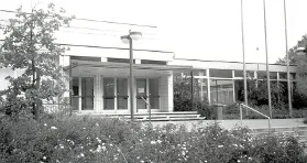 Schwarzweißfoto eines modernen Gebäudes mit Glasfront, Blumenbeet und Bäumen davor.