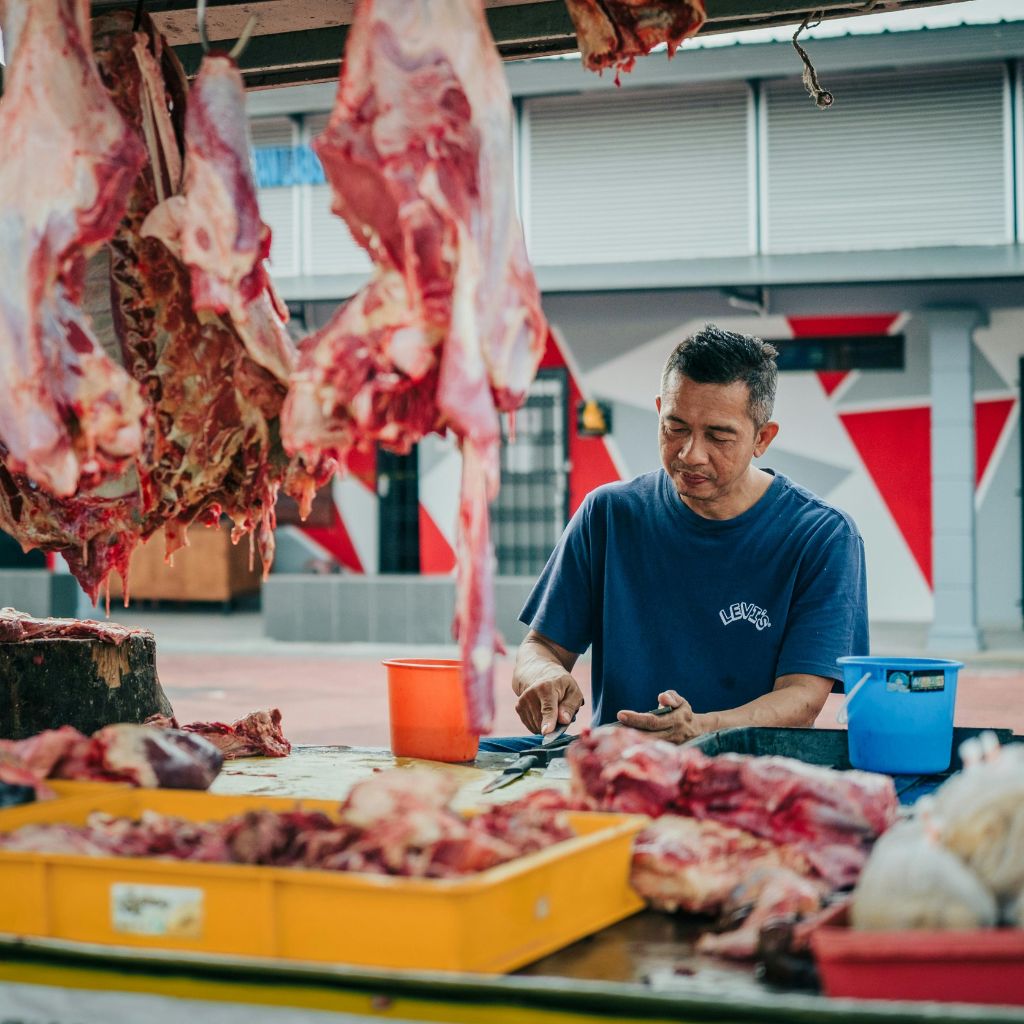 Man in blue shirt slicing raw meat at an outdoor market stall with hanging meat pieces.