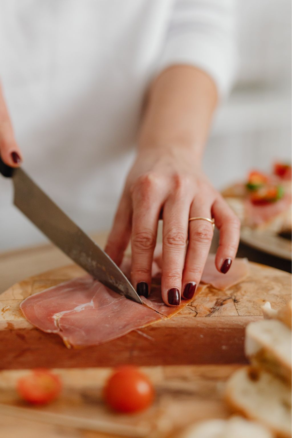 Person slicing thin prosciutto ham on a wooden cutting board with a kitchen knife.