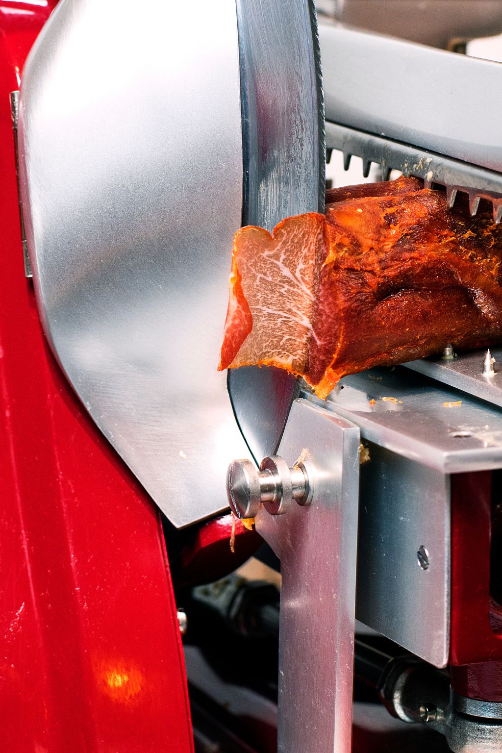 Red cured meat being sliced by a shiny metal meat slicer with a sharp circular blade.