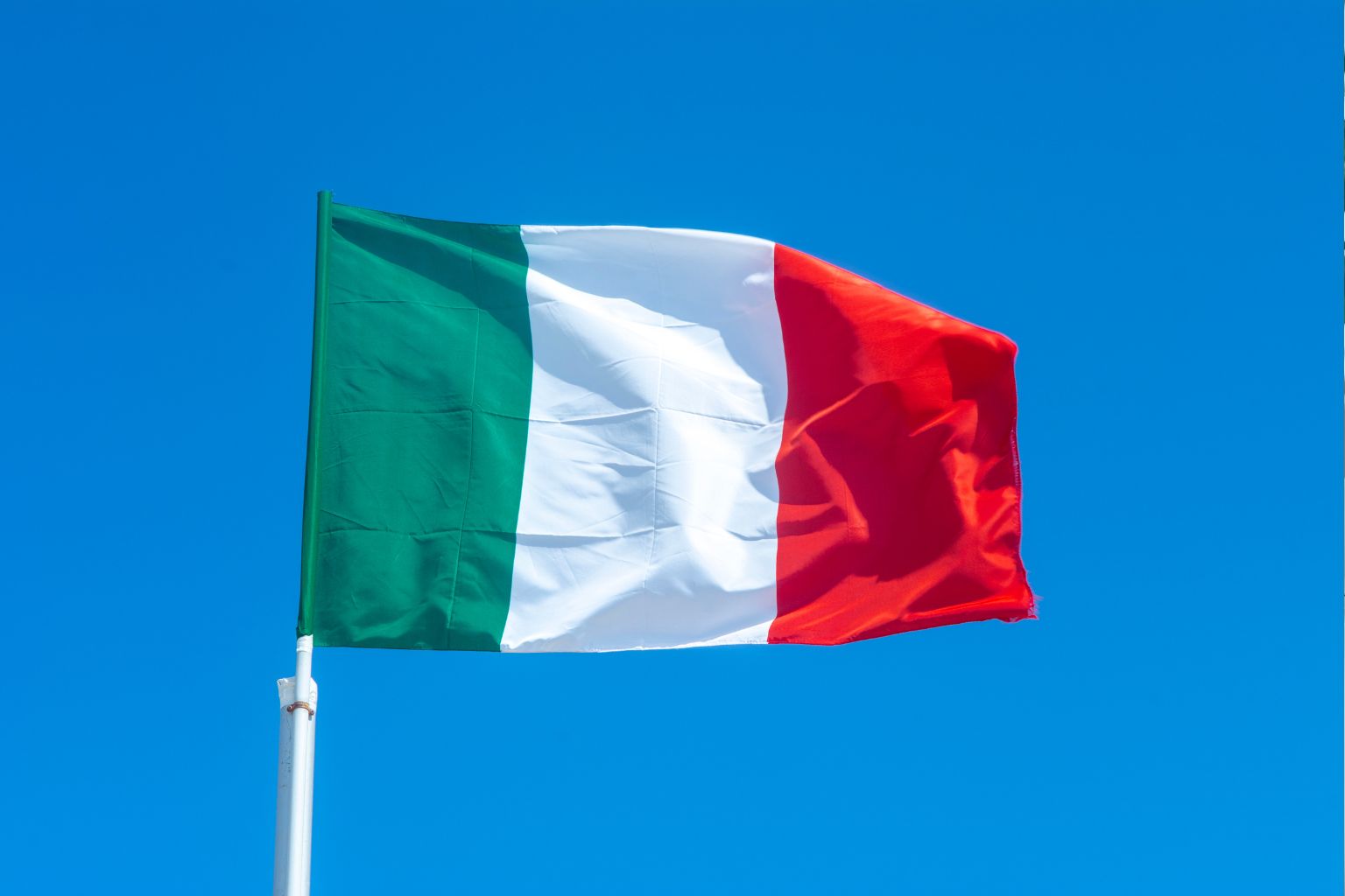 Italian flag with green, white, and red vertical stripes waving against a clear blue sky.