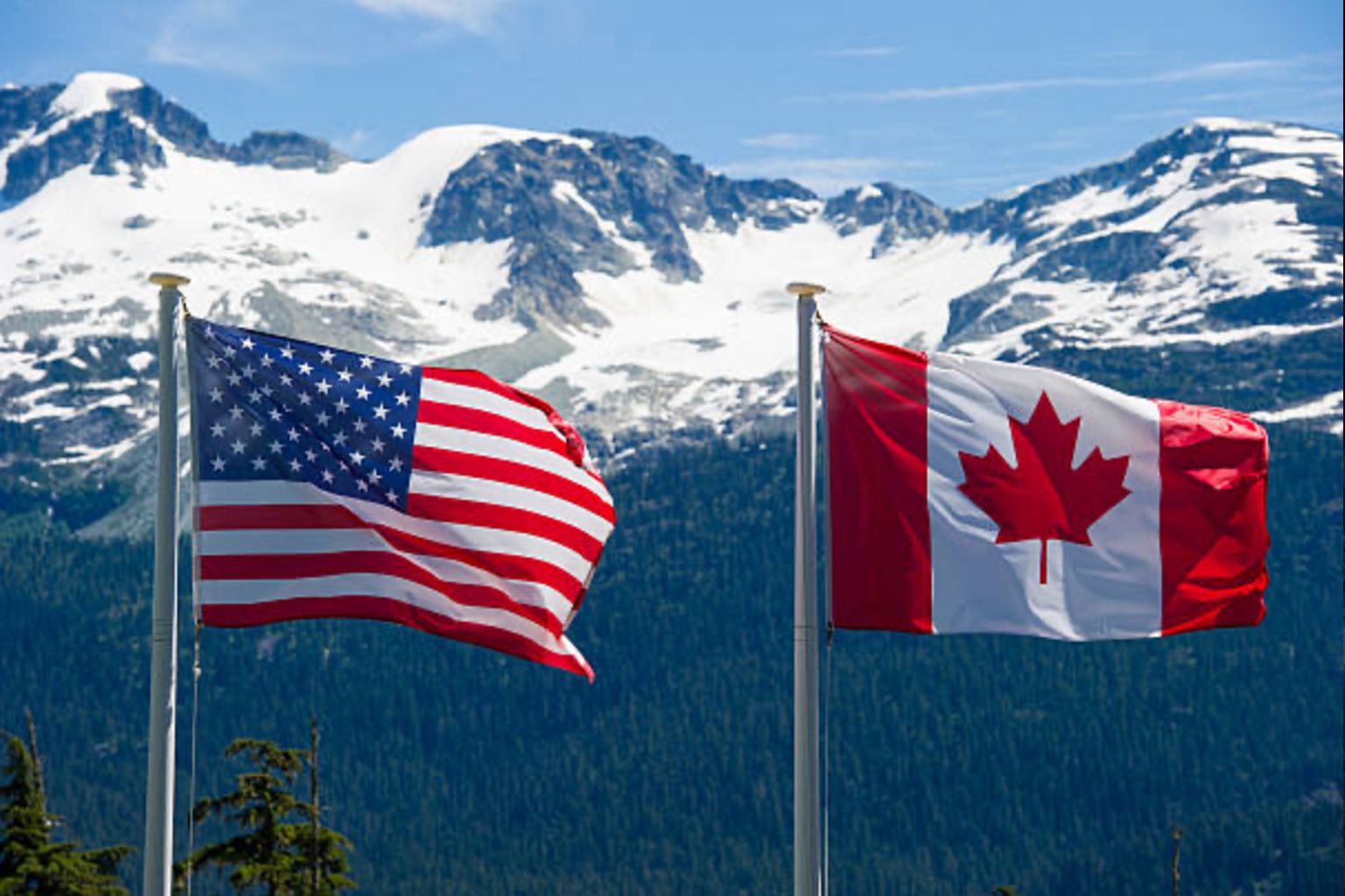 American and Canadian flags flying on poles with snow-capped mountains in the background.