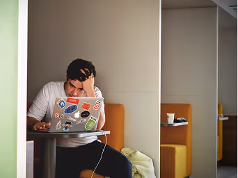 Man looking stressed while working on a laptop covered with stickers sitting in a cafe booth.