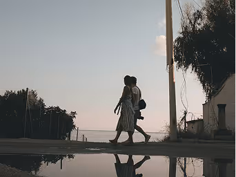 Couple walking outdoors near water at dusk with their reflection in a shallow puddle.