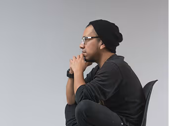 Man wearing glasses and a black beanie sitting thoughtfully with hands clasped and elbows on knees. Struggling with anxiety.