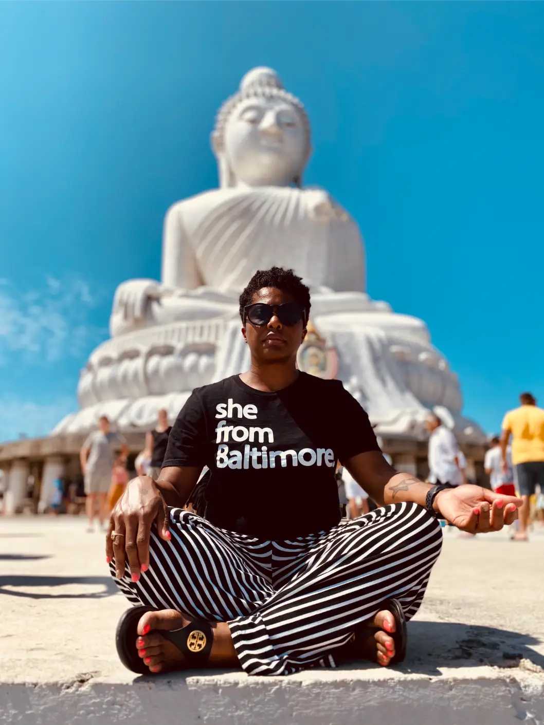 Person meditating cross-legged wearing striped pants and black shirt in front of large white Buddha statue.